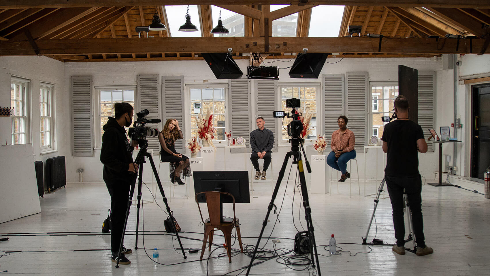 A shot from behind two cameramen filming a live streaming event in a large set with three presenters sat on stools