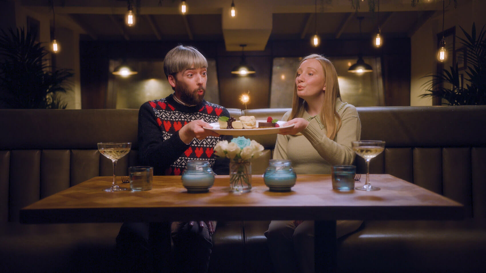 A couple in a restaurant on Valentines Day blowing out a sparkler on top of a dessert plate in a Buy A Gift ad, an example of branded content