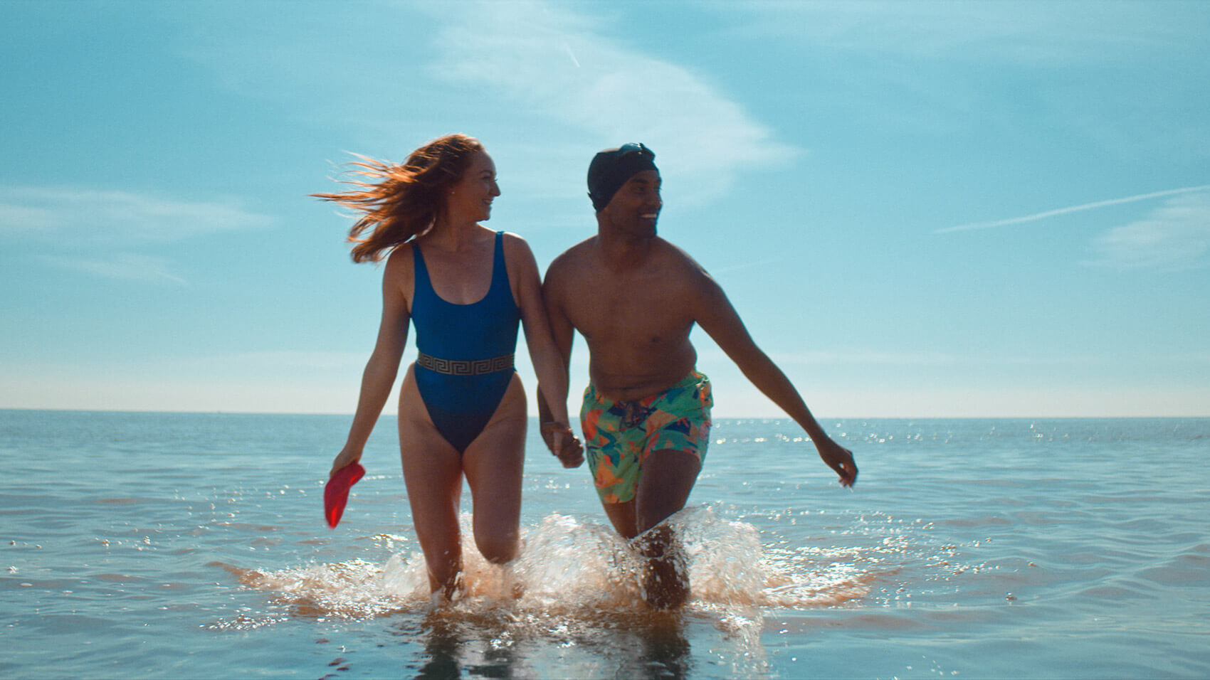 A female and male couple in swimming costumes smiling and holding hands as they run out of the sea in a branded content film for Hoo