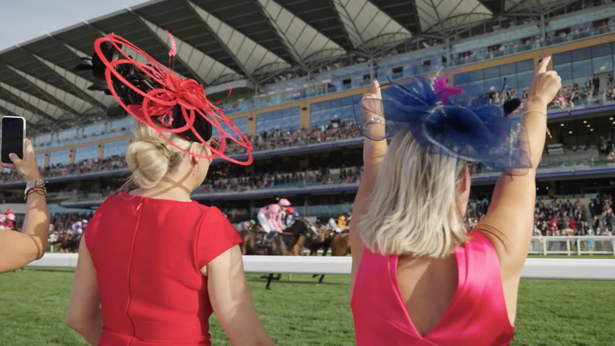 A still image of two spectators dressed up for Race Day watching a horse race