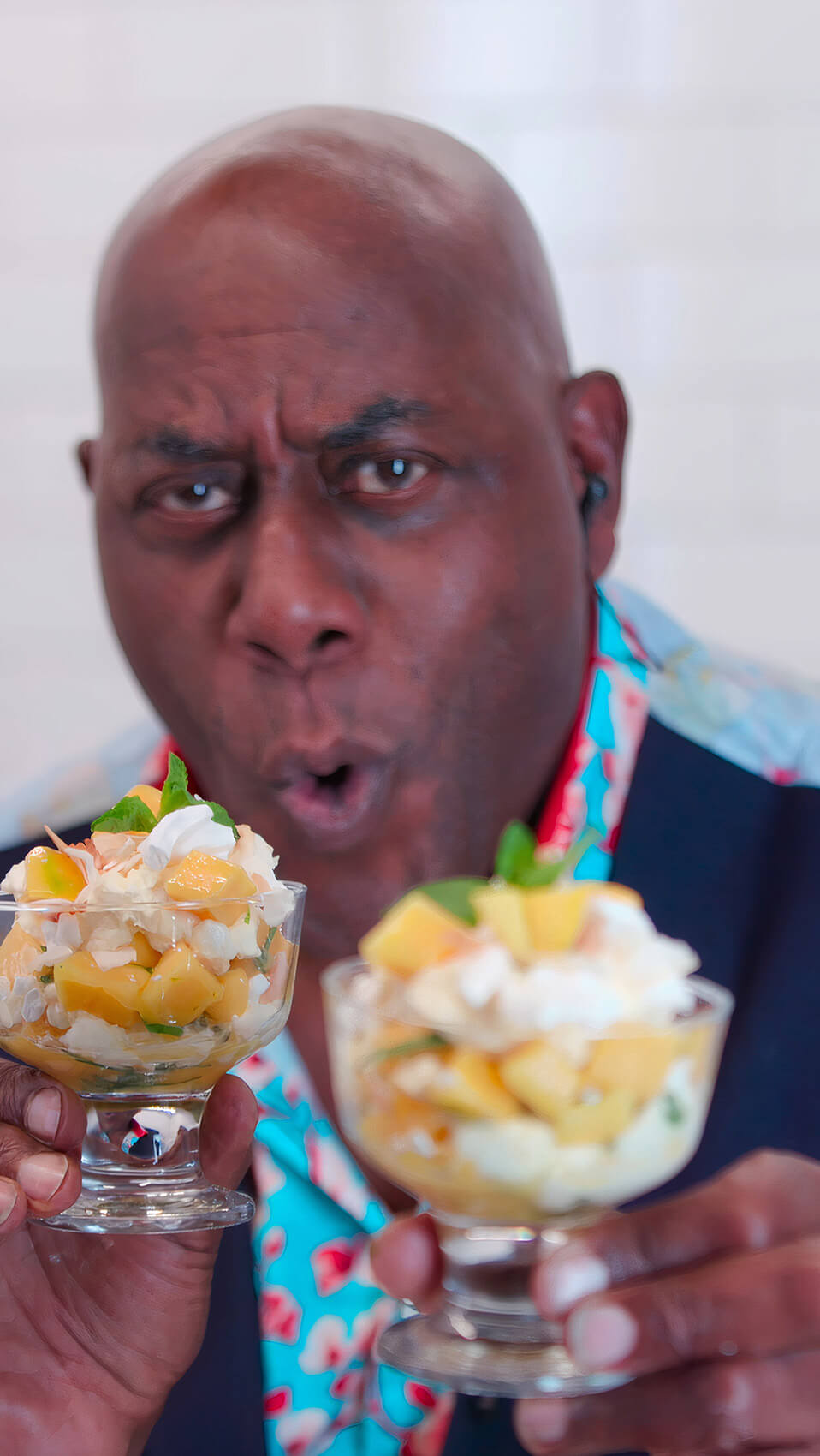 Ainsley Harriott holding two desserts in dishes during filming of a live streaming piece to camera