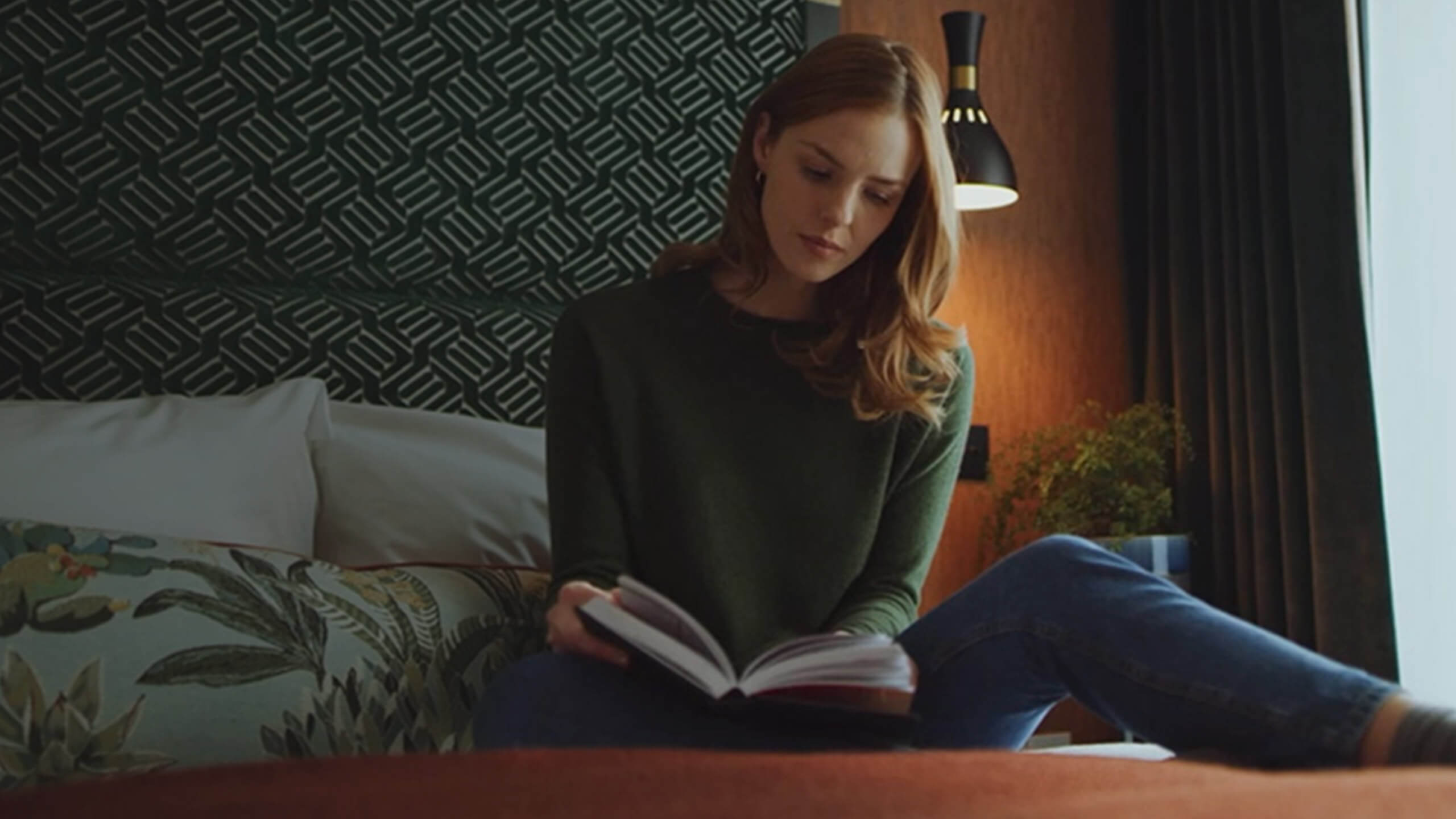 A still image from a property marketing video or property video production, of a woman sat on a bed reading a book in a modern room
